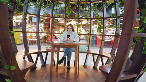 lady seated at decorative indoor table with leg crossed, looking at her phone, colorful decorative lights and greenery in background, providing a cozy atmosphere in the stylish sitting area