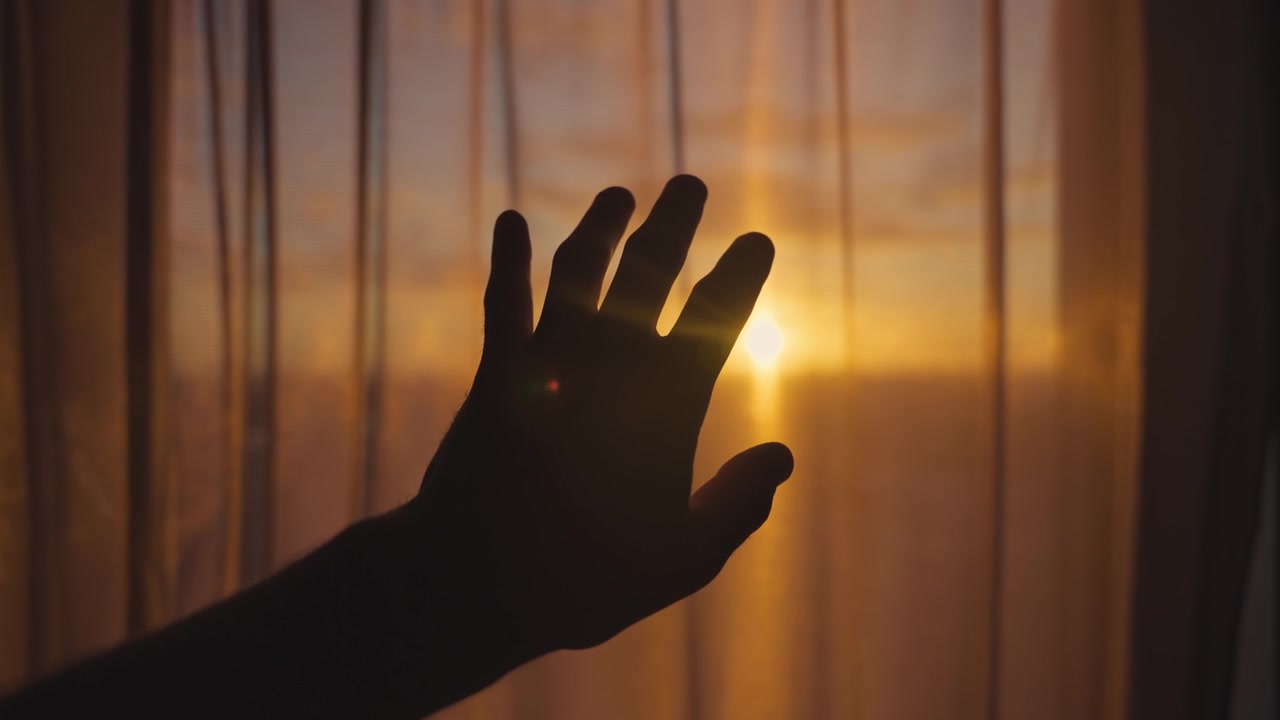 Silhouette of a man's hand moving against sunlit window and curtain inside a room. Sunset or sunrise as hand slowly blocking sun and moving around. Early morning warm sunlight. Hope and inspiration