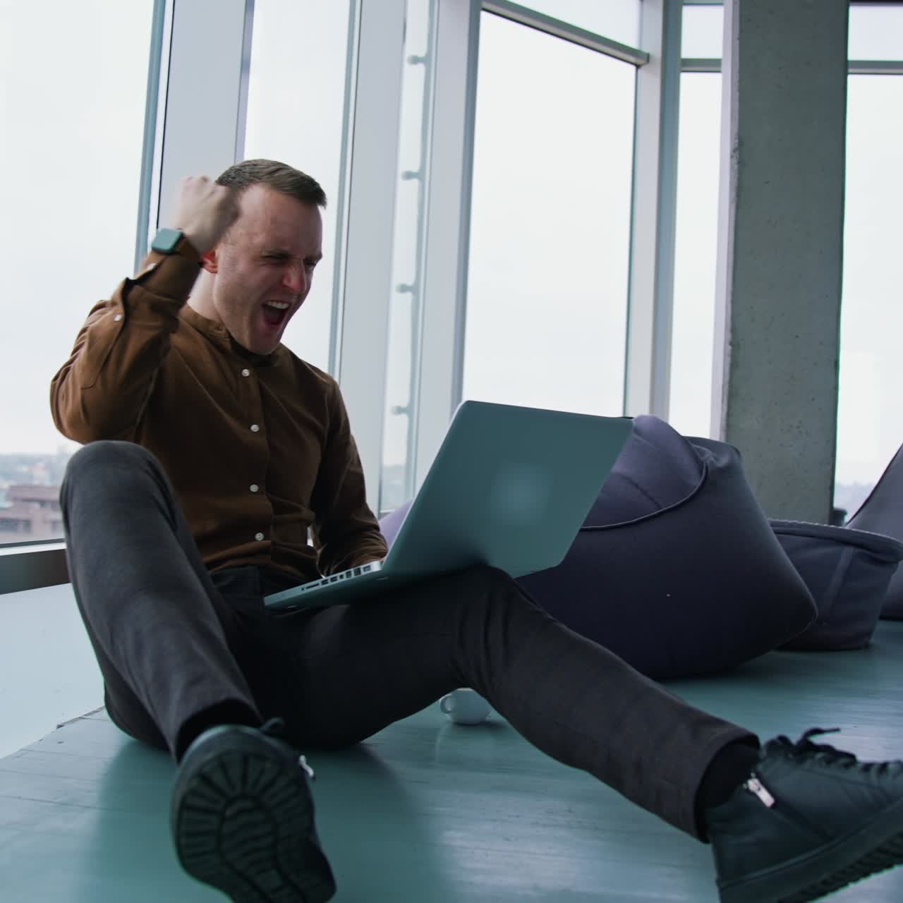 Young man sitting on floor near the window with a laptop. Happy man winning something while using a wireless computer