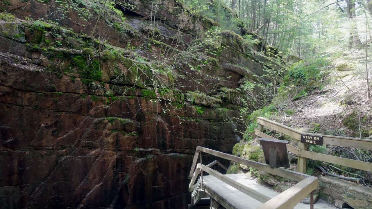 Visitors walk POV beside waterfall in narrow Flume Gorge path through red rock cliffs