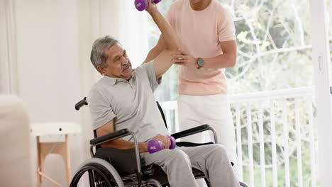 Diverse-male-physiotherapist-advising-and-senior-man-in-wheelchair-using-dumbbells,-in-slow-motion