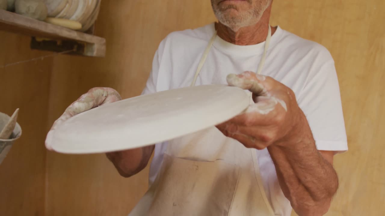 hombre caucásico mayor que usa un delantal inspeccionando un plato de arcilla en un taller de cerámica