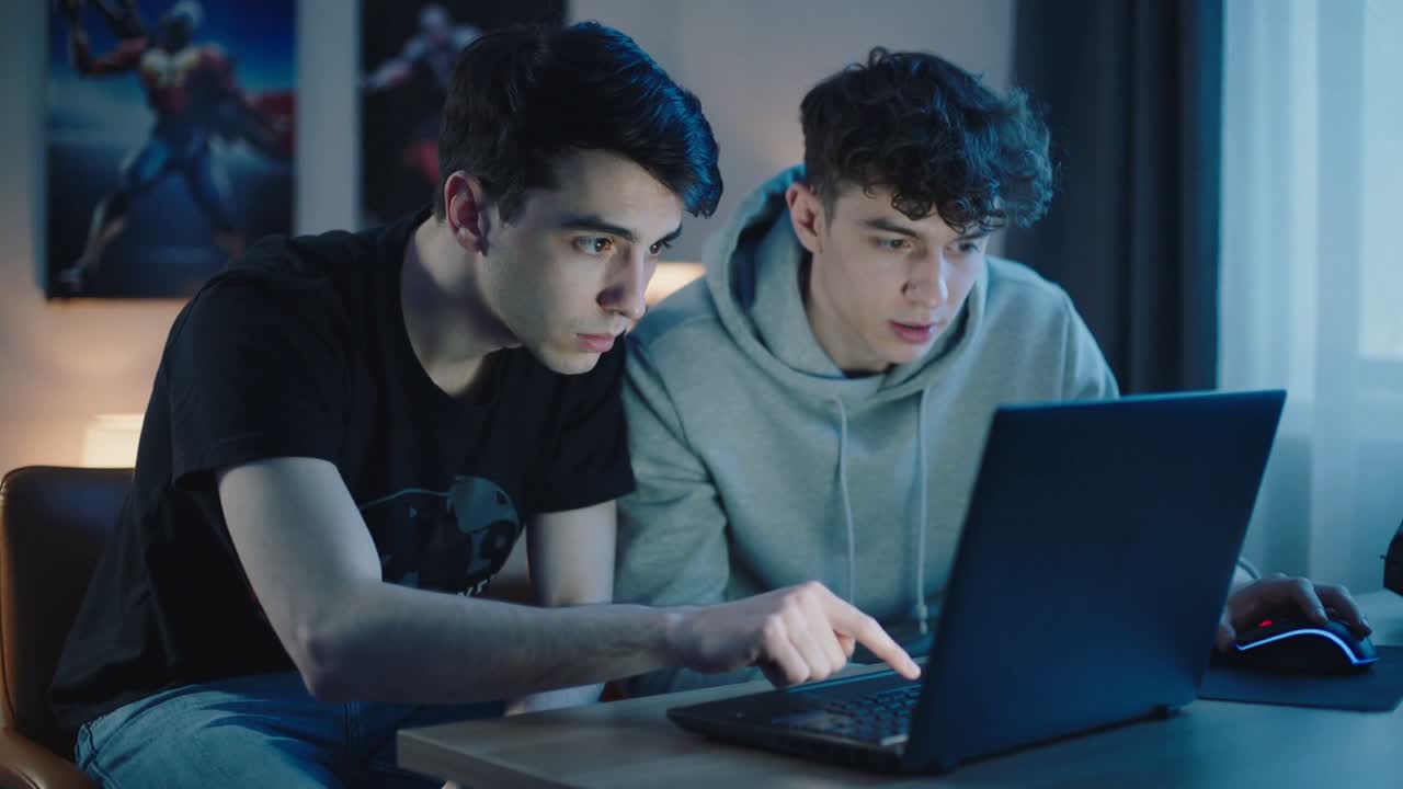 Pointing two teens examining laptop at home after alert, wearing graphic tee and gray hoodie, mouse