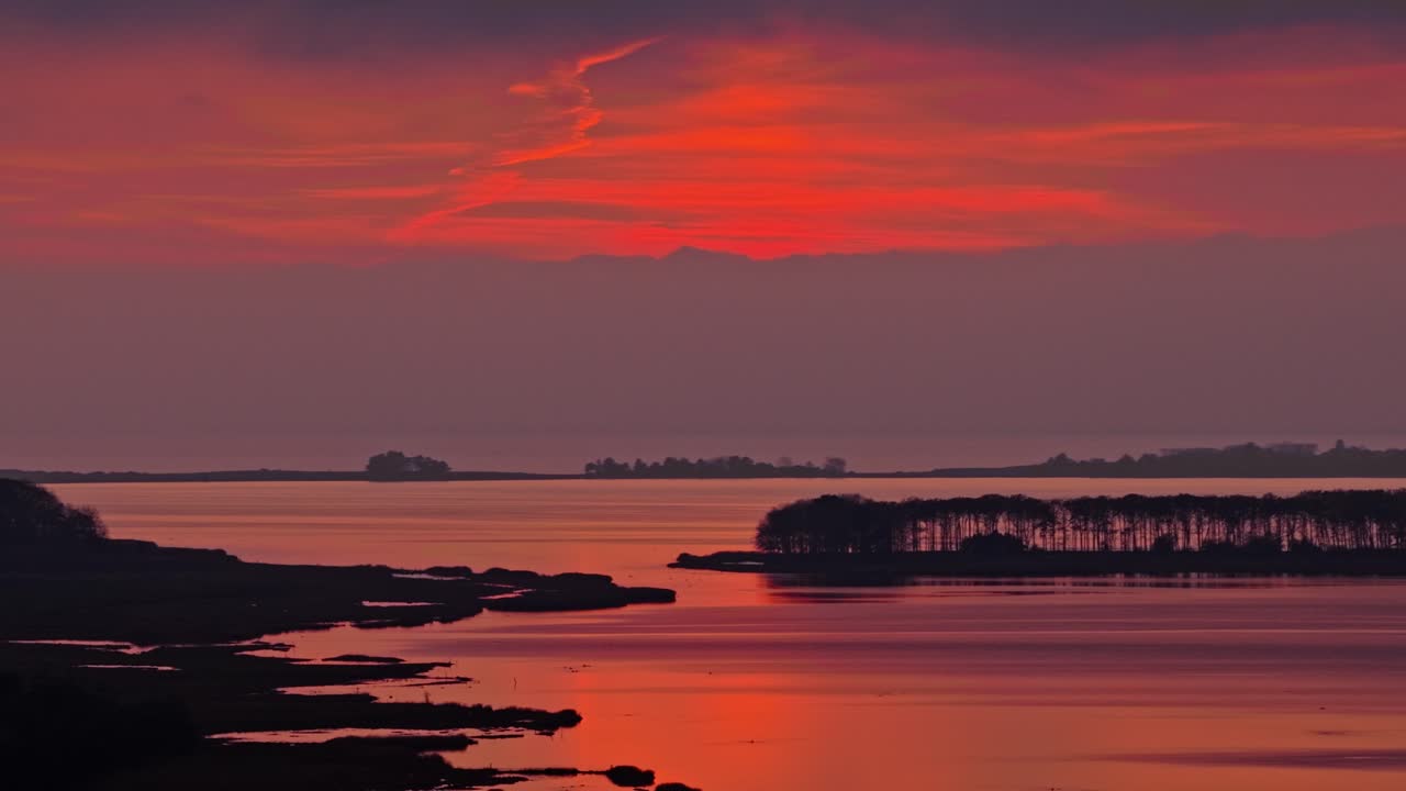 Slow pan revealing glowing red and orange sunset reflecting on still coastal water with dark tree silhouettes and distant islands. Peaceful nordic evening mood