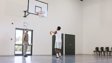 african american male basketball player catching, throwing basketball, male coach observing in gym