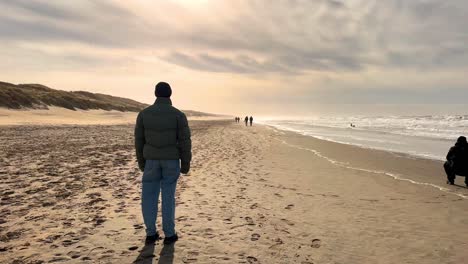 backwards shot of male person enjoying beautiful sunset at sandy beach with waves on sea during windy day