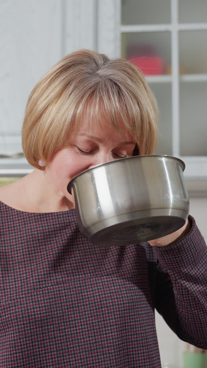 Mujer de mediana edad observando atentamente una olla que tiene en la mano con expresión concentrada en una cocina moderna; los armarios blancos del fondo crean un ambiente cálido mientras prepara la comida.