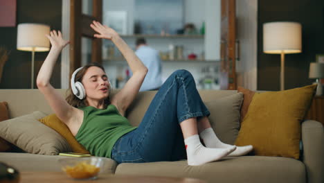 Mujer-Con-Auriculares-Disfrutando-De-La-Música-Poniendo-Un-Sofá-En-El-Interior.-Niña-Feliz-Moviendo-Las-Manos