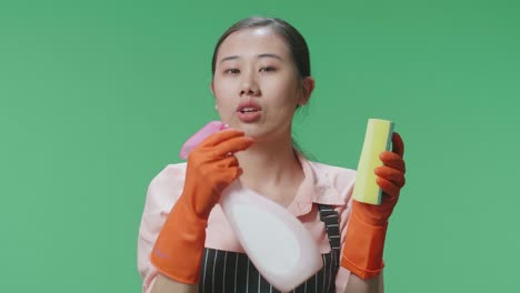 primer plano de una ama de casa asiática con un delantal sosteniendo una esponja de cocina y rociando antes de soplarla como un arma en el estudio de pantalla verde