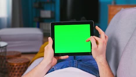 un joven en casa usa una tableta digital de pantalla verde. está tendido en un sofá en su acogedora sala de estar. un estudiante de sexo masculino se desplaza a través del contenido en la pantalla en blanco de su teléfono inteligente