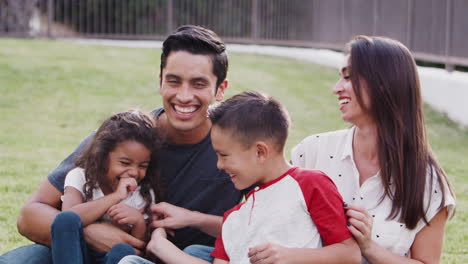 Feliz-Joven-Familia-Hispana-Sentada-En-El-Césped-Del-Parque-Haciéndose-Cosquillas-Y-Sonriendo-A-La-Cámara