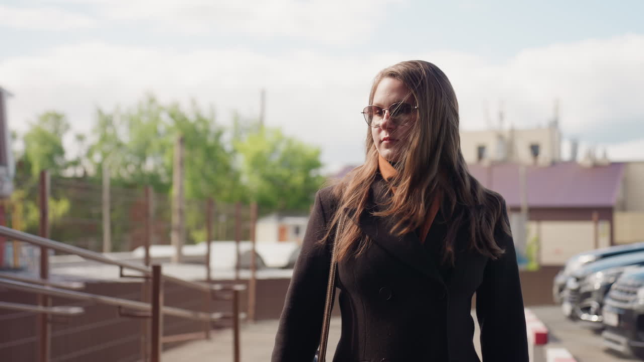 Un joven encantador con gafas de sol camina junto a una barandilla con una bufanda marrón sobre un abrigo oscuro mientras la brisa levanta su pelo, con un enfoque suave en las casas rojas detrás a la luz de la mañana, con un estado de ánimo tranquilo y un paso seguro.