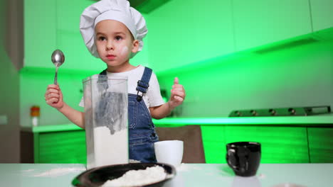 surprised baby boy in white chef cap stands at the table. kid with a spoon in hands in the kitchen. low angle view.