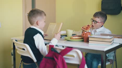 un escolar come pizza y un amigo lee un libro de texto en la mesa.