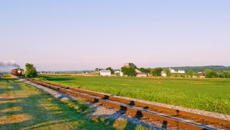A train moves along railway tracks surrounded by lush fields and distant farmhouses during a colorful sunset. The vibrant sky adds to the tranquil rural atmosphere of the moment