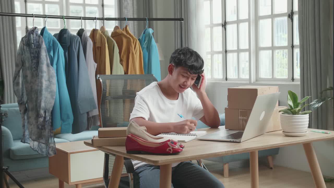 Asian Man Online Seller Talk On Mobile Phone And Take Note While Using Computer For Selling Clothes At Home