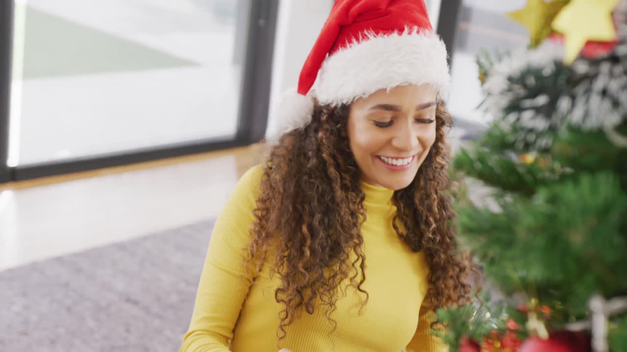 video de una feliz mujer biracial en el sombrero de santa sonriendo y decorando el árbol de navidad en casa