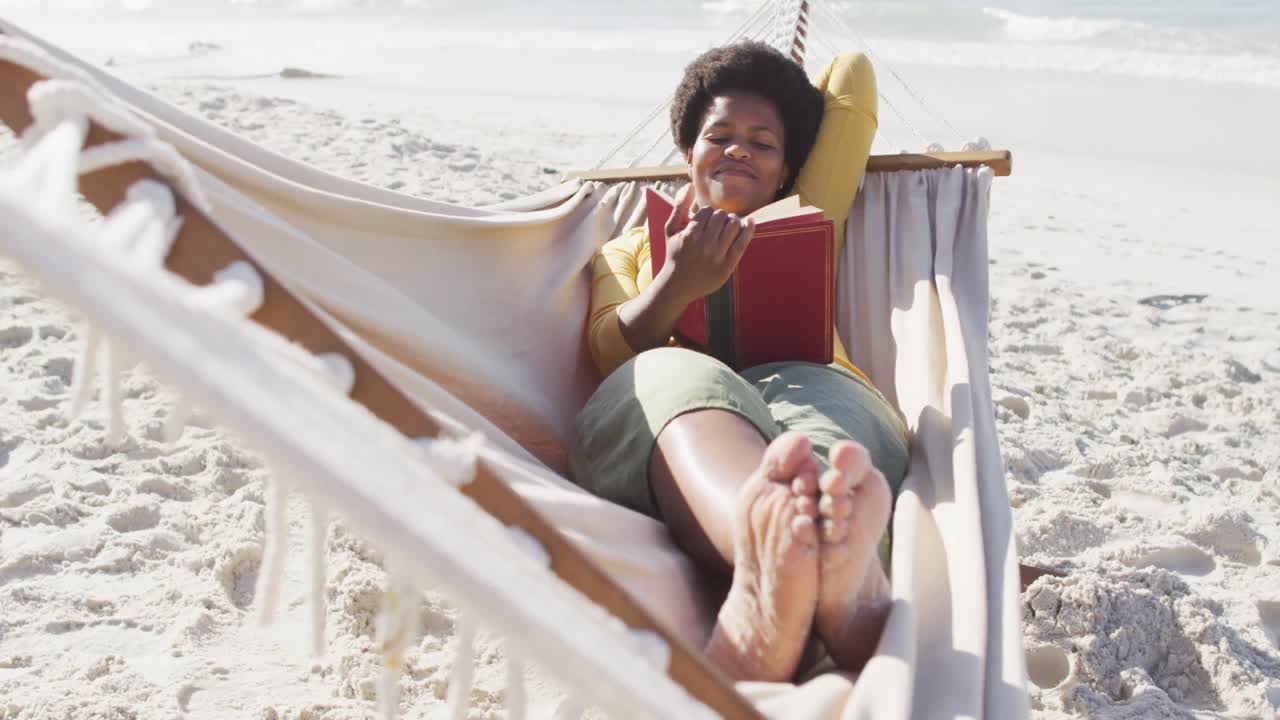 feliz mujer afroamericana leyendo y acostada en una hamaca en una playa soleada