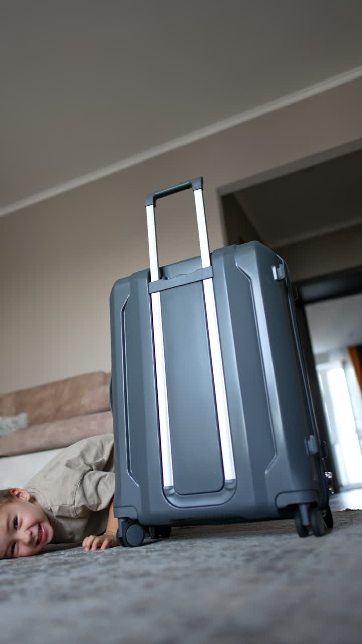 Funny cheerful boy hides behind the big grey suitcase. Smiling kid crawls by the floor. Vertical video.