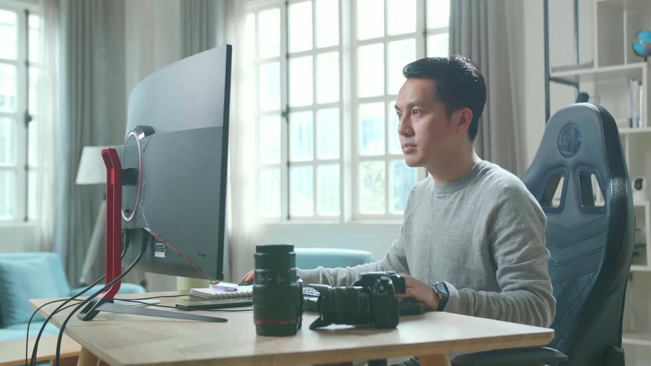 Asian Cameraman In Long Sleeved T-Shirt Using Desktop Computer On A Table For Working At Home.