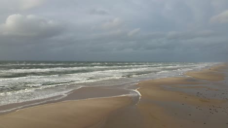 a medium-altitude truck shot starts over the endless beach at hvide sande in denmark, slowly moving towards the rough ocean, capturing the expansive sandy landscape and turbulent sea on a stormy day