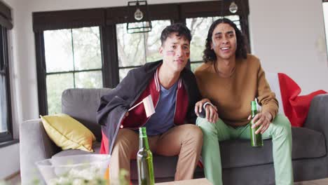 Happy-diverse-male-couple-watching-tv-with-national-flags-and-supporting