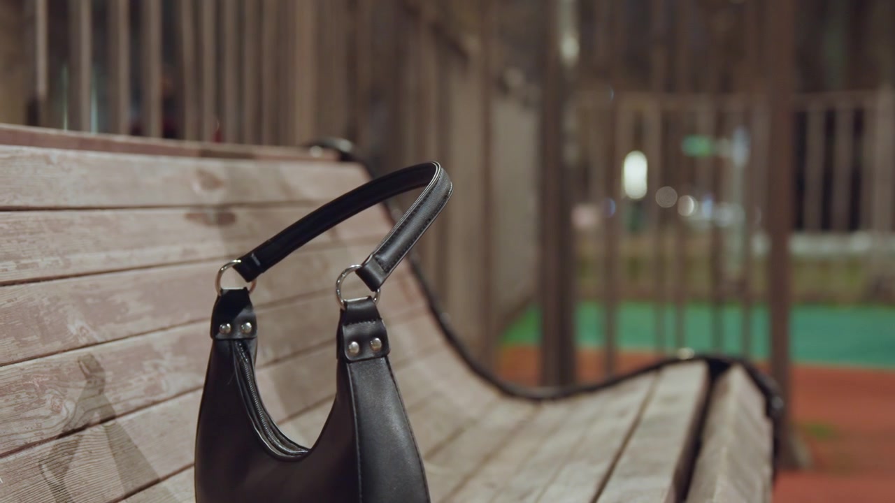 夜、ベンチに革製ハンドバッグを置く白人女性のクローズアップ。チェーンストラップと金属製の金具に焦点が当たり、隣には空席が見える。木の質感とランプの光が、一時停止とわずかな緊張感を醸し出している。