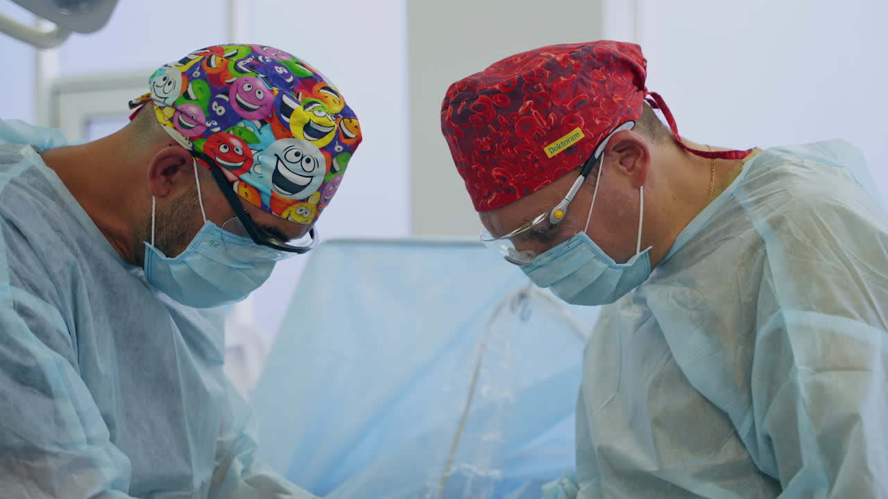 Surgeons in funny caps bent over the patient at surgery. Doctors work hard standing on both sides of a patient.
