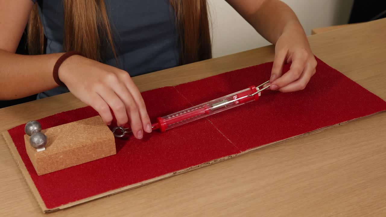 Hands use a spring scale to measure friction on sandpaper with a weighted block. Bright lighting highlights the setup