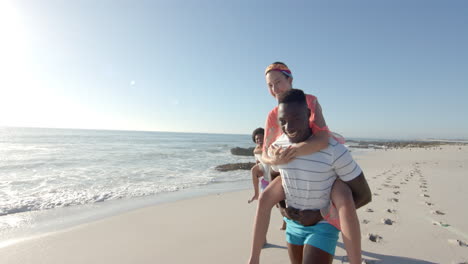 ein junger afroamerikanischer mann fährt auf dem rücken einer jungen kaukasischen frau am strand