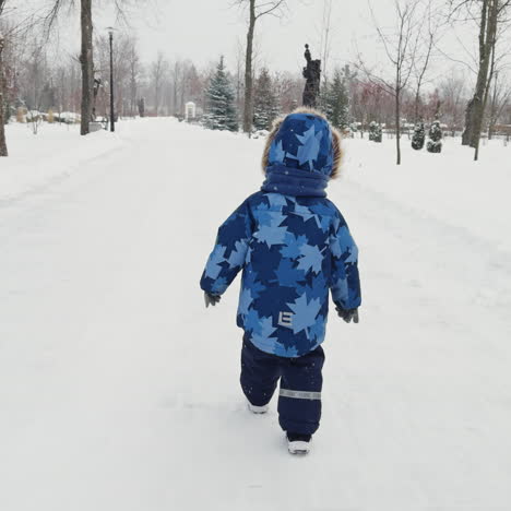 following the little kid wearing warm clothes walking by the winter park. toddler outdoors on snowing weather.