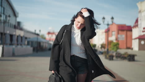 lady in black coat, short leather pant and white sweater adjusts her hair back, carrying a black handbag, with sun reflecting off her face in an urban setting