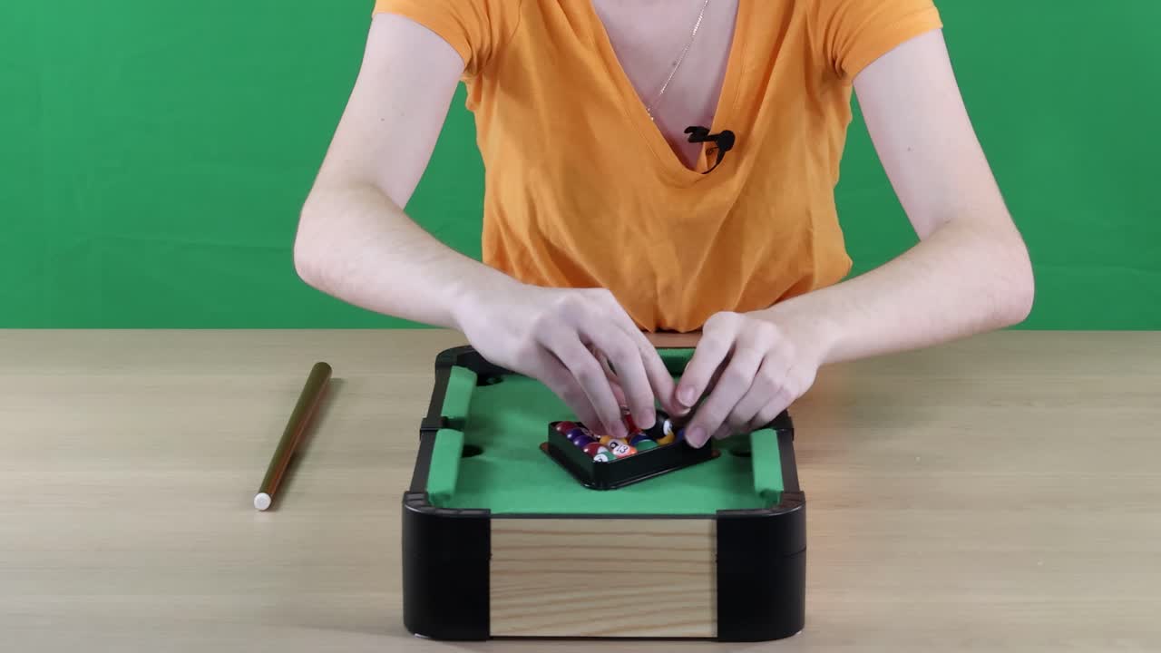 A person arranges balls on a mini pool table with a cue stick nearby, set against a green screen backdrop