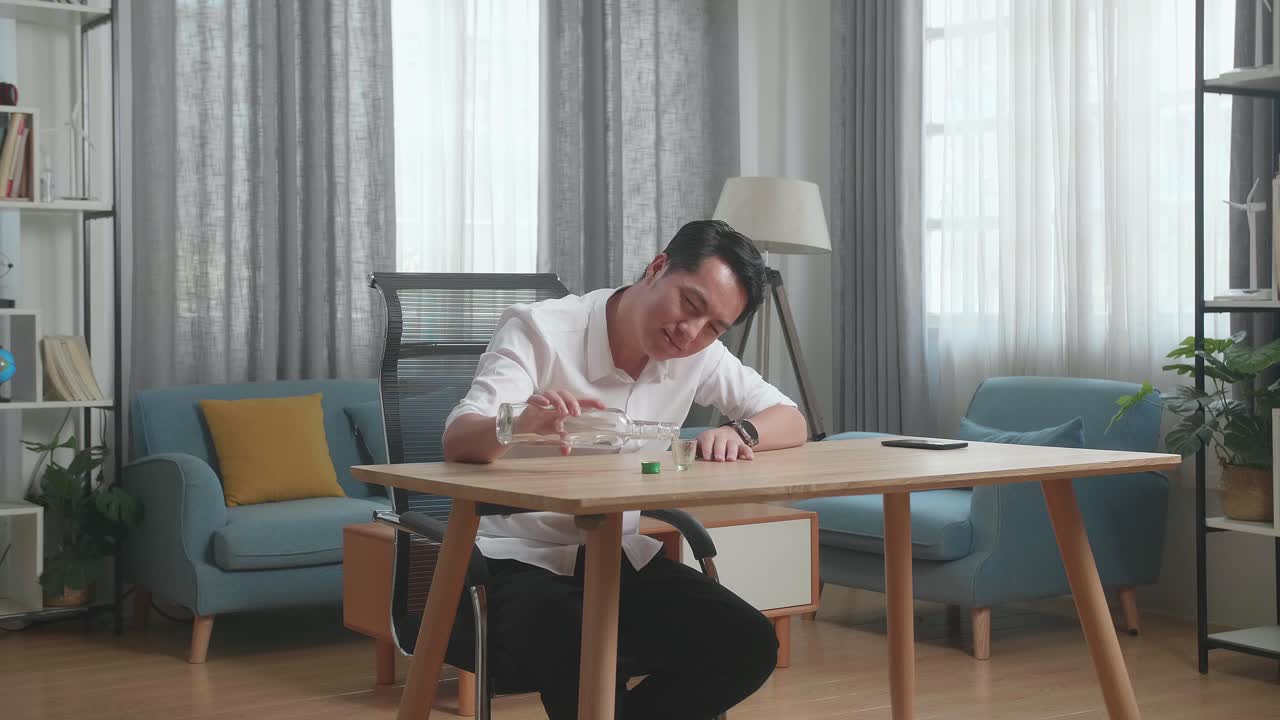 술에 취한 아시아인 남자가 테이블 위에 스마트폰을 들고 미소 짓고 집에서 술을 마시기 전에 크 컵에 보드카를 부었습니다.