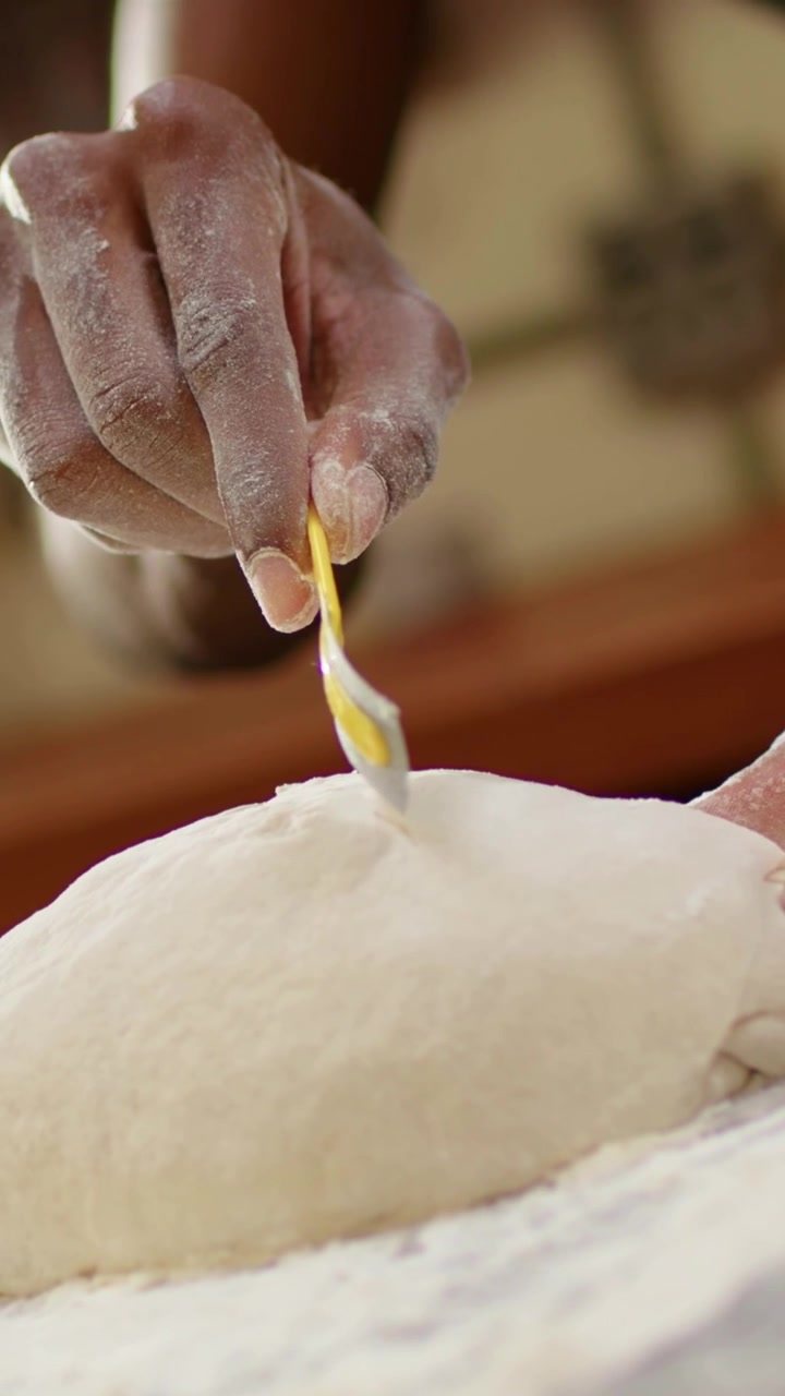 Video verticale: Panettiere con grembiule che incide l'impasto in cucina con uno strumento giallo, modellando una pagnotta.