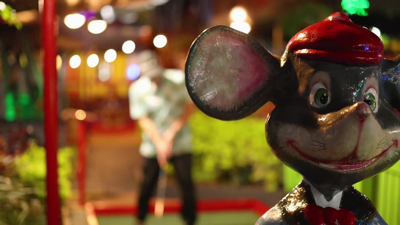 A nighttime miniature golf scene with a mouse statue and a player preparing to putt under colorful lights