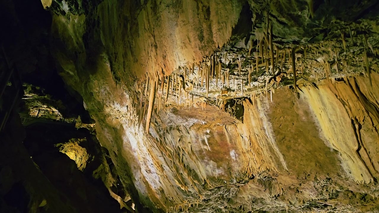 Scenic cave chamber features towering stalagmites, long stalactites, rugged boulders, and colorful blue and yellow lights creating a dramatic subterranean landscape for tourism footage.