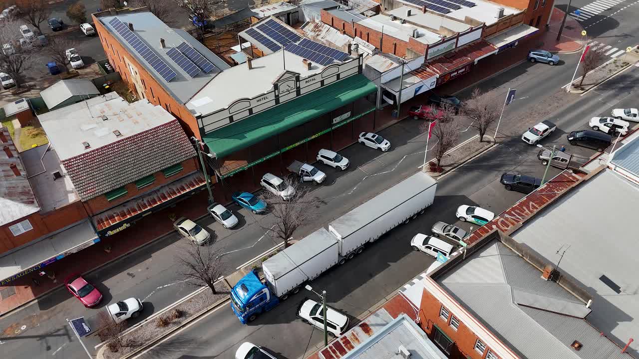 A large semi truck with a blue cab slowly drives through a narrow urban street lined with parked cars and low-rise buildings under daylight