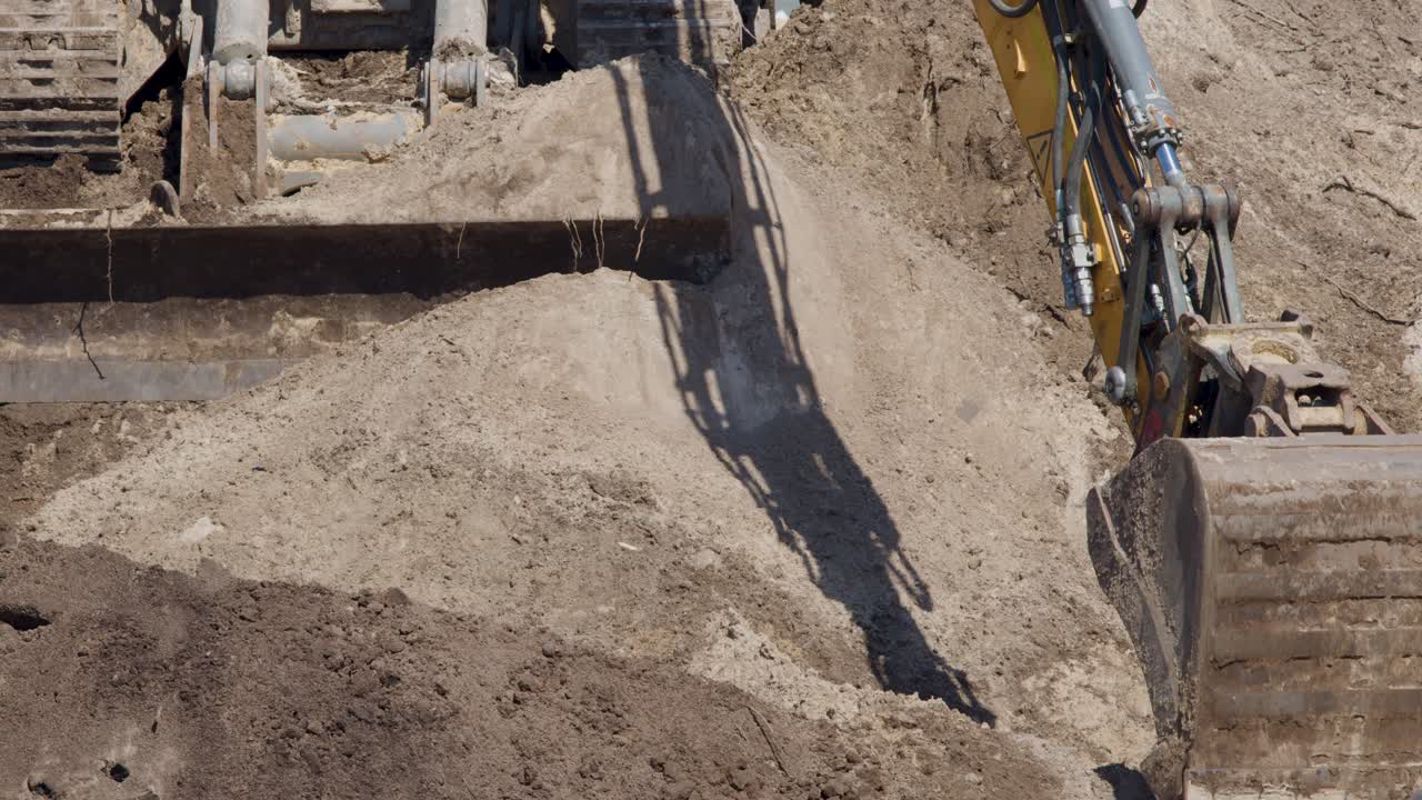 Excavator releases soil from bucket onto ground, bright daylight, static overhead camera, construction environment