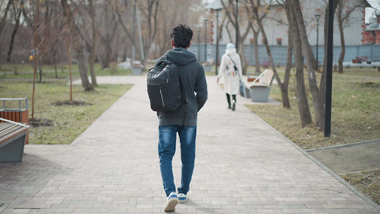 Man in casual clothing with backpack walking alone along paved park path on overcast day, trees lining walkway, distant woman ahead, relaxed atmosphere suggesting daily routine or personal journey