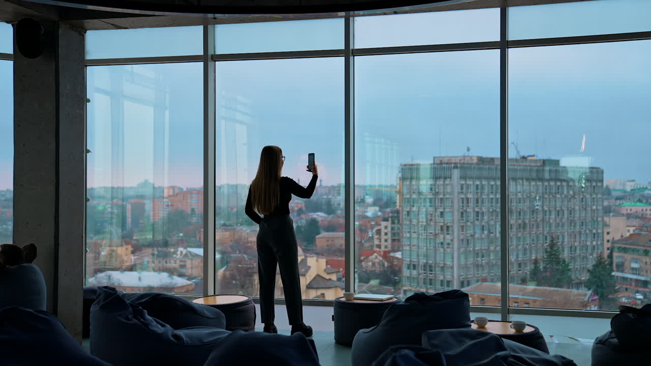 Young woman with a phone near window. Panoramic office view with large windows on city background. Rear view of a girl indoors photographing the evening city landscape.