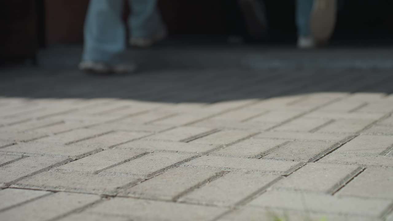 Legs in sneakers walking along interlocked pavement path under sunlight, creating dynamic shadows on ground, suggesting casual outdoor activity or journey on urban pedestrian walkway