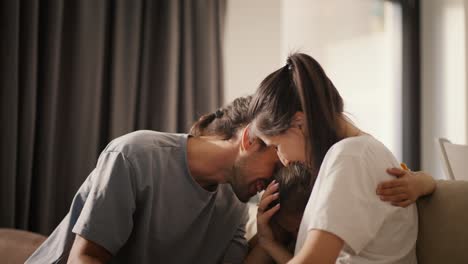 Close-up-shot-of-a-brunette-man-in-a-gray-T-shirt-and-his-wife,-a-brunette-girl-in-a-white-T-shirt,-hugging-their-little-daughter,-a-brunette-girl-in-a-yellow-dress-who-jumps-on-the-sofa-and-hugs-her-parents-in-a-cozy-modern-room