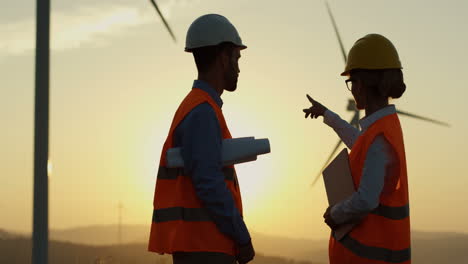 kaukasische ingenieurinnen und ingenieure in helm und uniform unterhalten sich, während sie etwas über den betrieb der windmühlen an der station für erneuerbare energien entscheiden