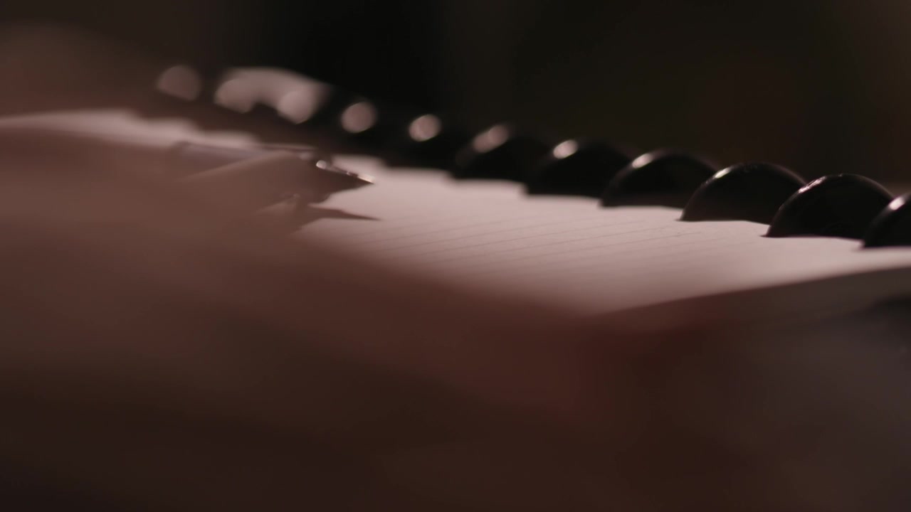 Dita che scrivono su una tastiera in primo piano sfocato con una penna e un blocco note a fuoco sullo sfondo.
