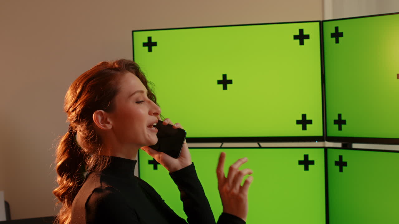 Young woman talking on phone, multiple green screen computer monitors with tracking