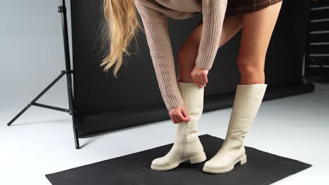 Blond long haired lady bends to check her elegant light high boot. Model footage of stylish modern footwear on plain soles and low heel.