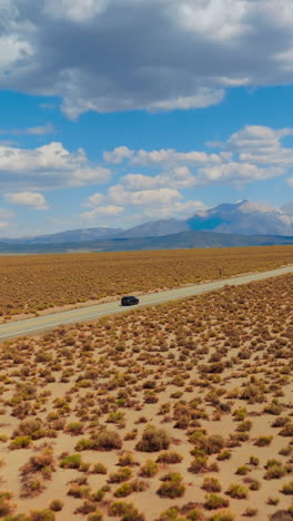 yellow dry deserted landscape contrasting with bright azure sky with multiple clouds. black car riding by the road. mountains at backdrop. vertical video