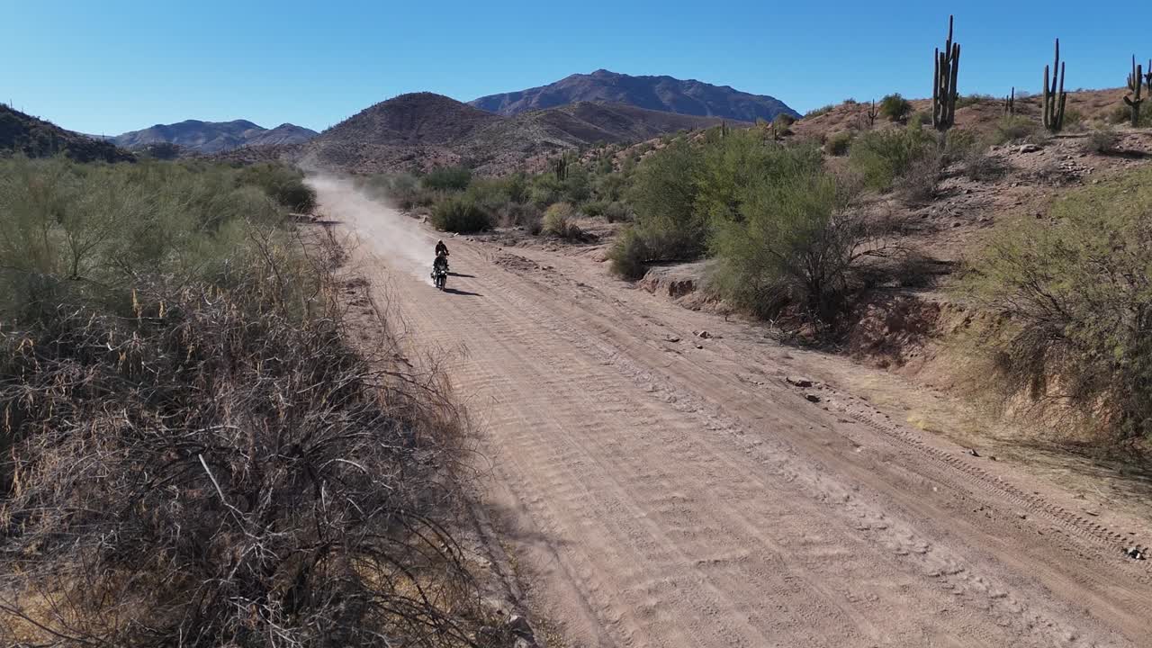 Dirt Bikes Riding down a Desert Road, kicking up dust behind them. Mountains Brush and Cactus in the background. Arizona