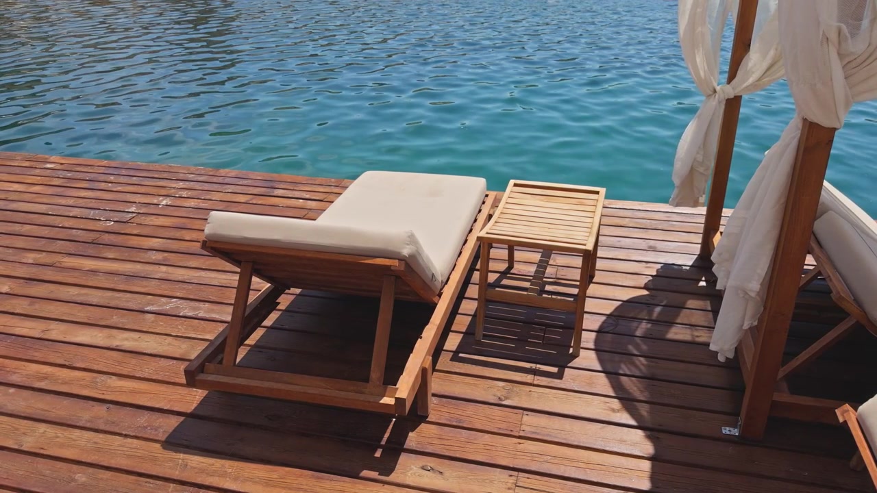 Wide shot of empty sun lounger and side table on sunny wooden dock overlooking bright blue sea, luxury beach club setting, Mediterranean coast, Turkey.